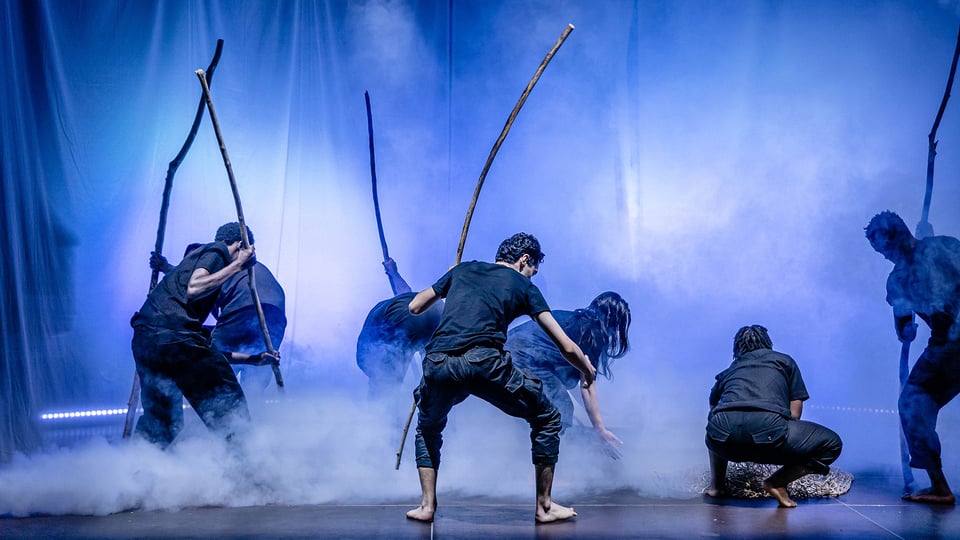 Bühnenfoto aus Mut! Theater grenzenlos mit mehreren Darstellenden in schwarzer Kleidung auf einer nebelgefüllten Bühne, die lange Äste in den Händen halten und vor blau beleuchteter Kulisse in unterschiedlichen Positionen agieren