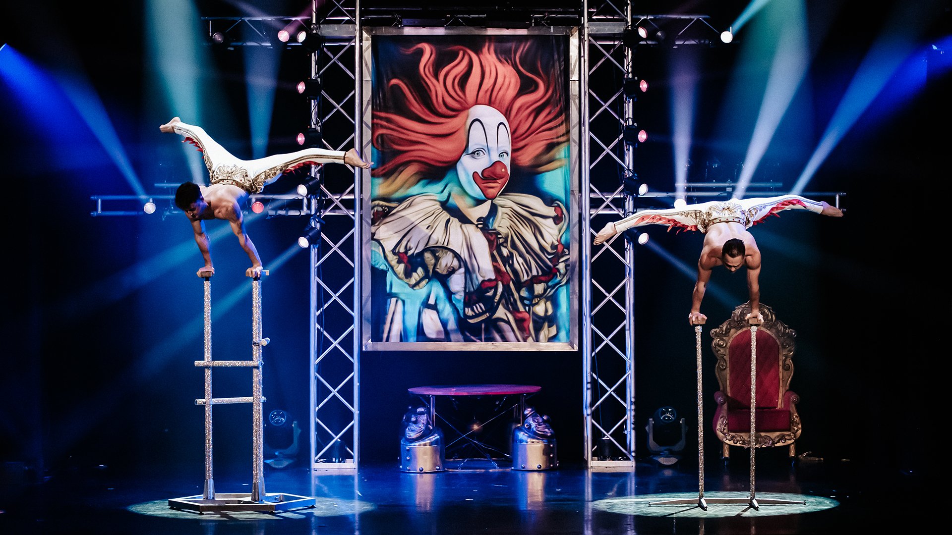 Bühnenfoto aus der GOP-Show Circus mit zwei Artisten im Handstand auf hohen Handstandstäben, die waagerecht über den Geräten balancieren, vor einem großen Clownsmotiv und blau ausgeleuchteter Bühnenkulisse