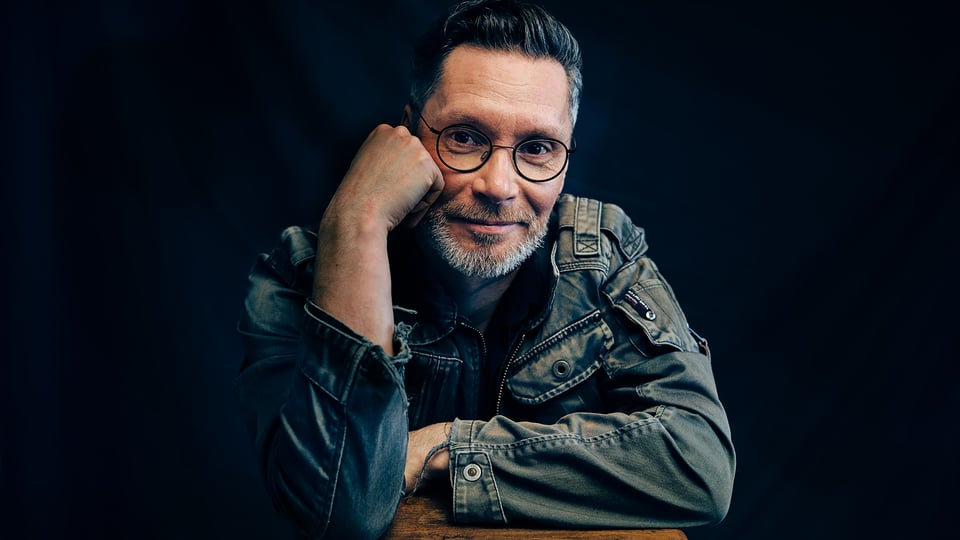 Zur Veranstalrtung Staubkind - eine Person in Jeansjacke und mit Brille, den Kopf auf eine Hand gestützt vor dunklem Hintergrund