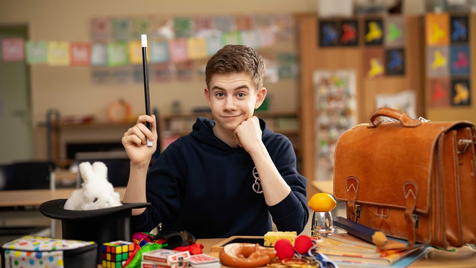 Foto von Magic Maxl an einem Tisch in einem Klassenraum mit Zauberstab, Zylinder, Spielkarten, Rubikwürfel, Zauberrequisiten und brauner Lederschultasche im Vordergrund.