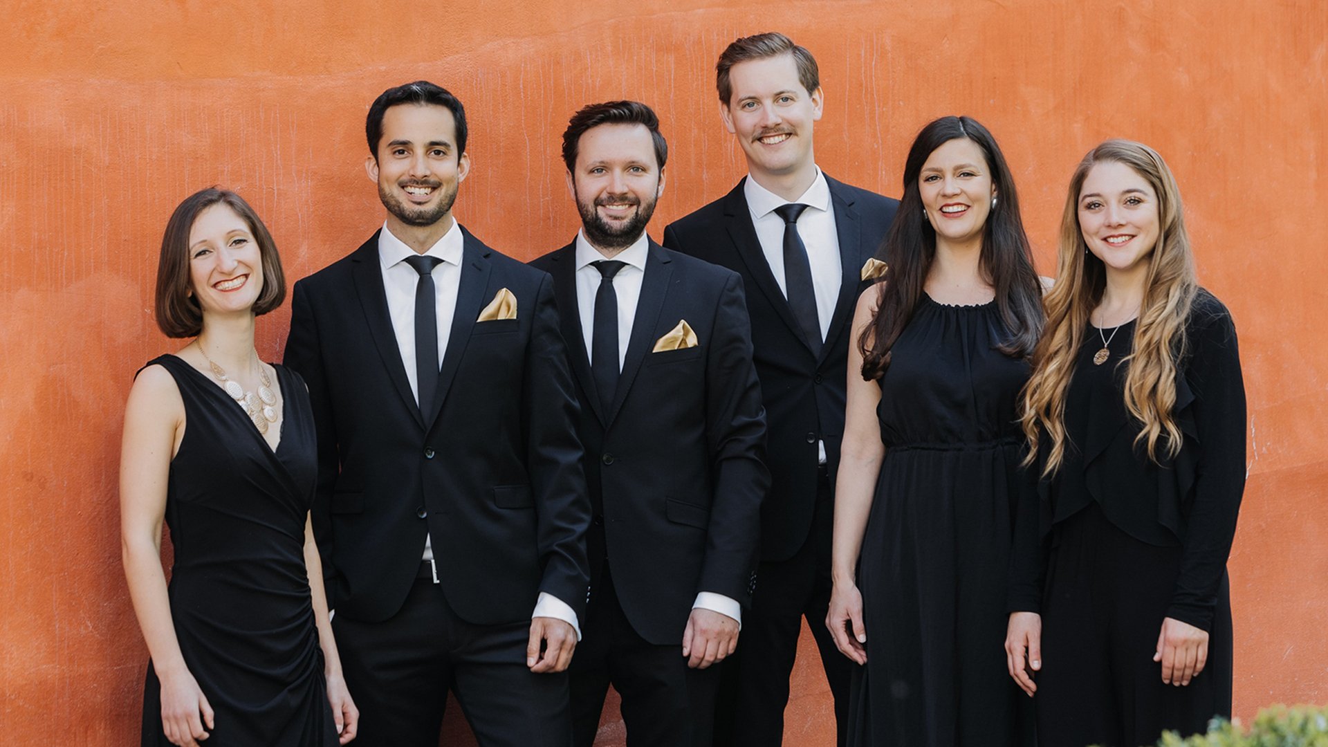 sechs Personen in dunkler Kleidung vor orangefarbenem Hintergrund 