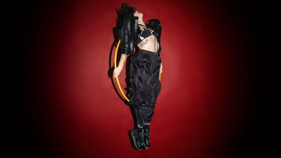 Foto für die Veranstaltung „MINE – Tour 2026“ mit der Künstlerin in Schwarz mit Hula-Hoop vor rotem Hintergrund.