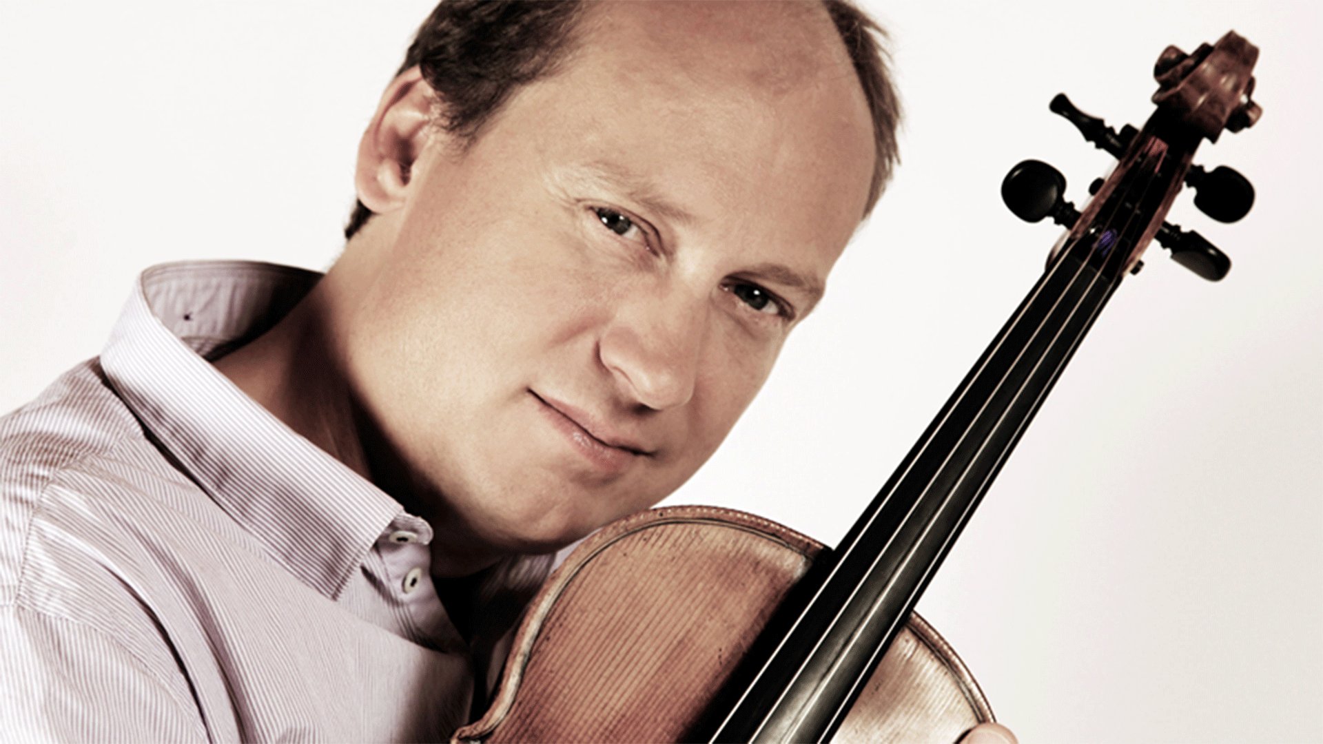 Nahaufnahme einer Person mit Violine vor hellem Hintergrund. Das Instrument wird an die Schulter gehalten, während die Person direkt in die Kamera blickt.