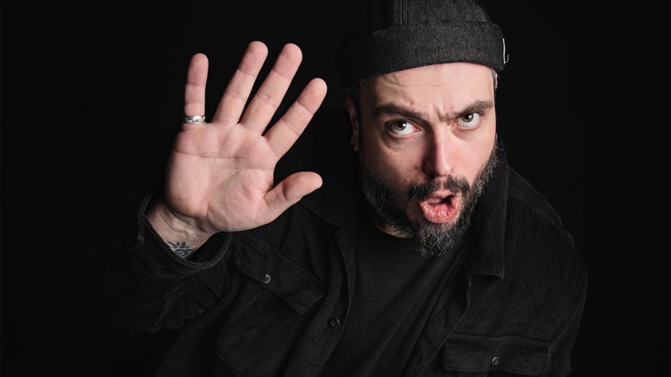 Eine Person in dunkler Kleidung hebt eine geöffnete Handfläche Richtung Kamera; die Szene ist vor schwarzem Hintergrund in dunkler, kontrastreicher Beleuchtung aufgenommen.