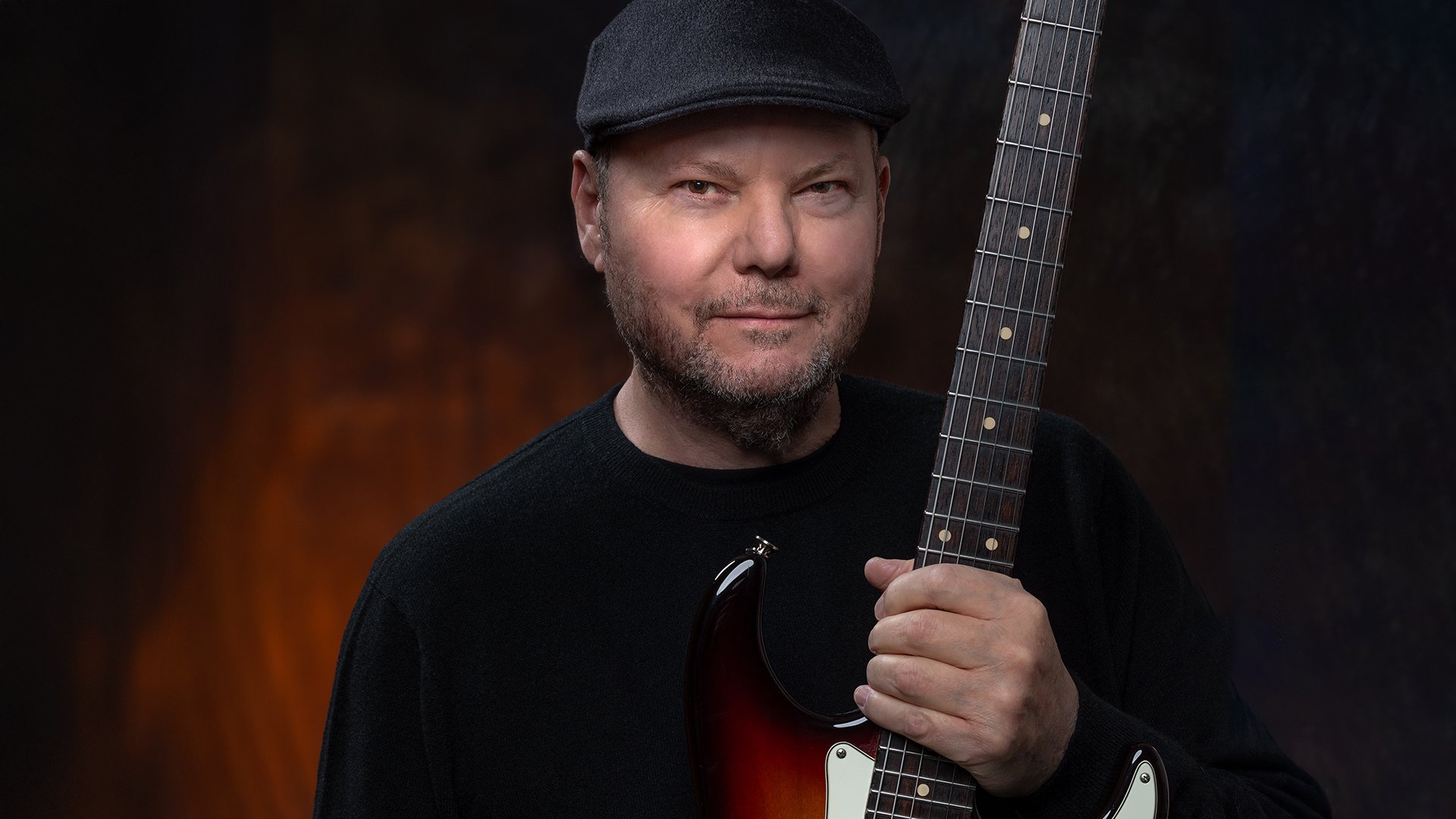Christopher Cross mit Schiebermütze und Gitarre vor braun marmoriertem Hintergrund