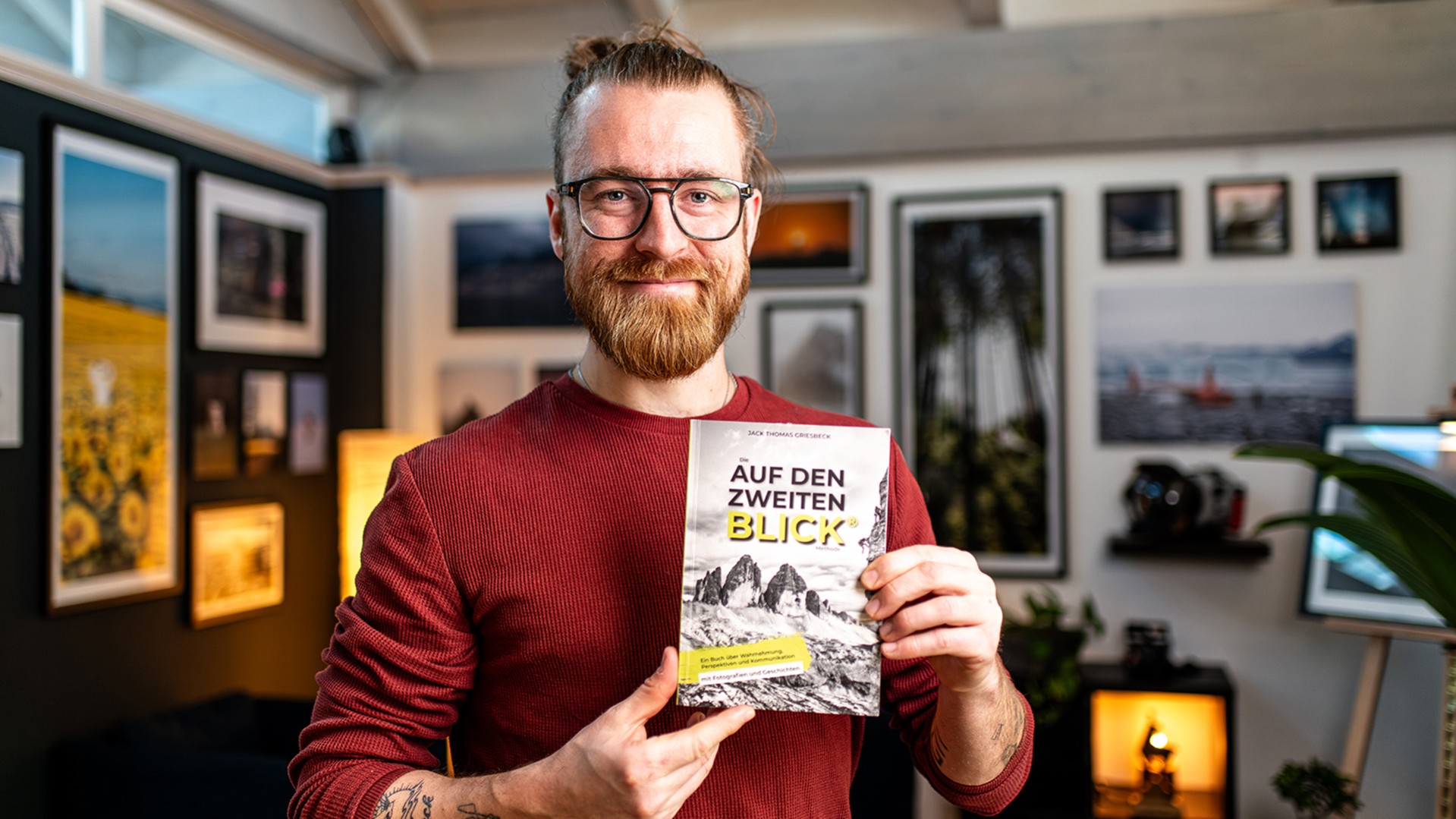 Ein Mann mit Brille und rotem Pulli hält einen Flyer mit dem Titel auf den zweiten Blick in der Hand, im Hintergrundmehrere gerahmte Bilder an der Wand
