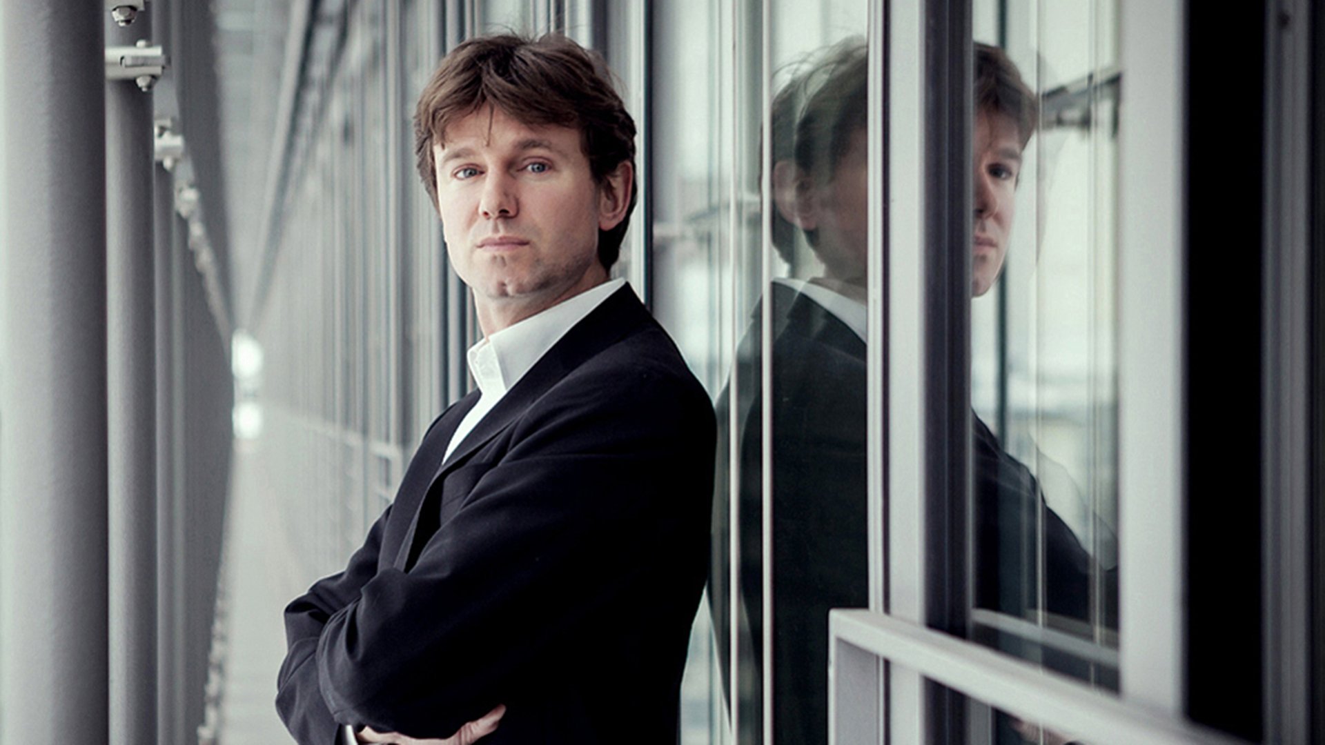 Ein Künstler in dunklem Anzug an einer spiegelnden Fassade lehnend 