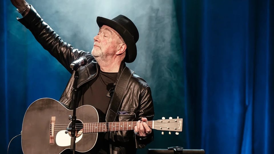 Mann mit Ledejacke, Hut und Gitarre auf Bühne, im Hintergrund blauer Vorhang 
