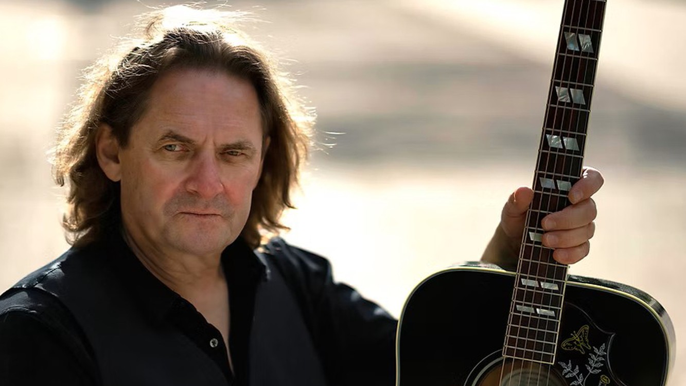 Porträt von Rusty Stone im Freien, die eine schwarze Akustikgitarre am Hals hält, heller, unscharfer Hintergrund.