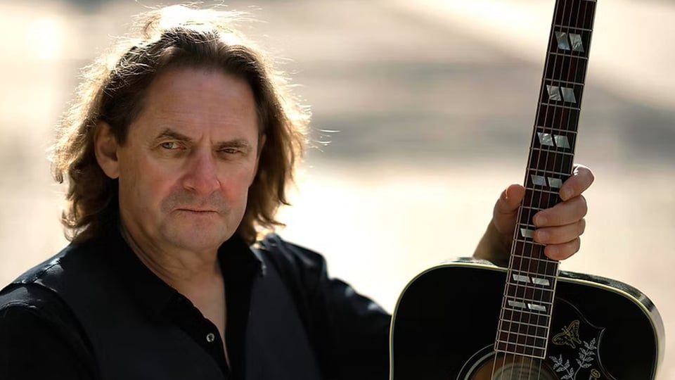 Porträt von Rusty Stone im Freien, die eine schwarze Akustikgitarre am Hals hält, heller, unscharfer Hintergrund.