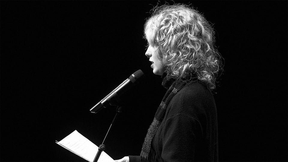 Schwarz-weiß Aufnahme einer Person mit lockigem Haar am Mikrofon, liest von einem Blatt auf einer Bühne – Poetry Slam oder Lesung mit starkem Kontrast.