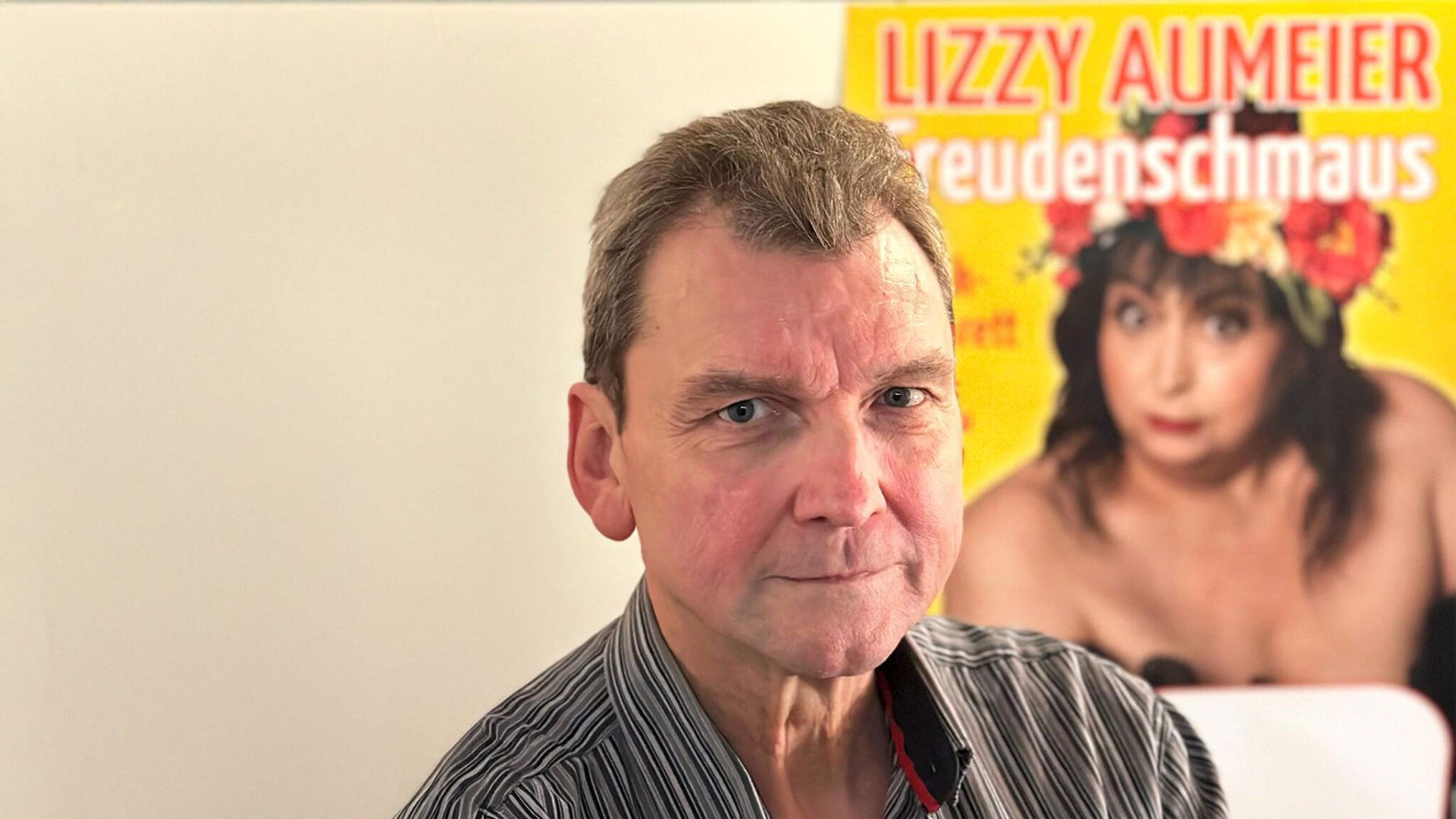 Foto für das Event von Andreas Stock mit dem Künstler und im Hintergrund einem Poster der verstorbenen Lizzy Aumeier