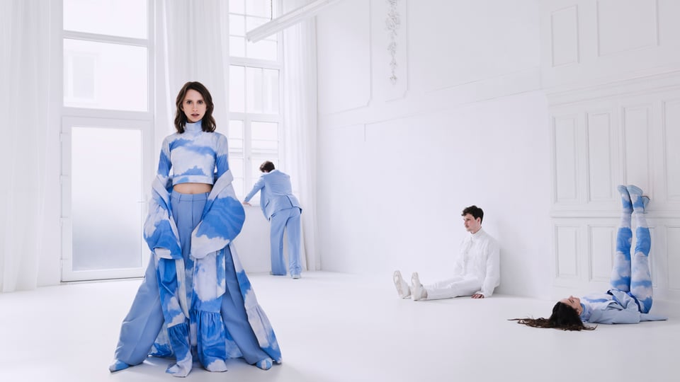 vier Personen in weißer und blauer Kleidung, eine Dame im Vordergrund, in weißem Raum mit hohen Wänden