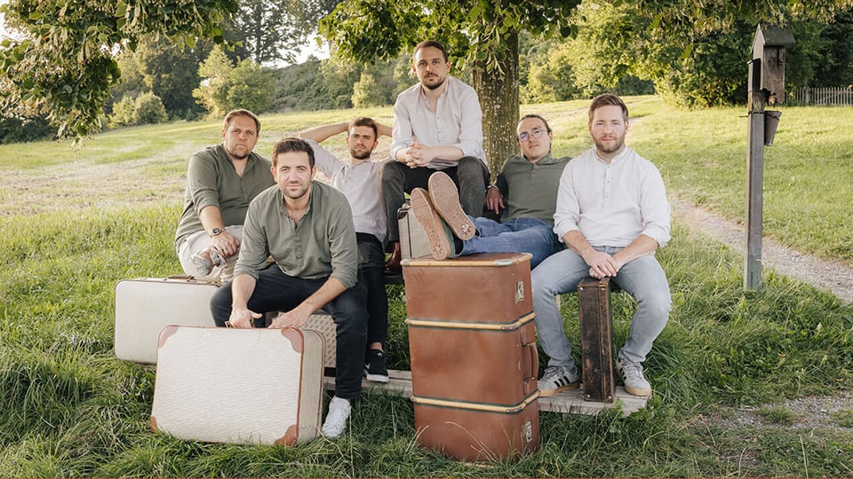 Eine Gruppe von sechs Personen sitzt entspannt auf einer Holzbank unter einem Baum im Grünen. Vor ihnen stehen mehrere große, vintage‑artige Reisekoffer. Die Szene wirkt ländlich und sommerlich, mit Wiese, Bäumen und einem Weg im Hintergrund.
