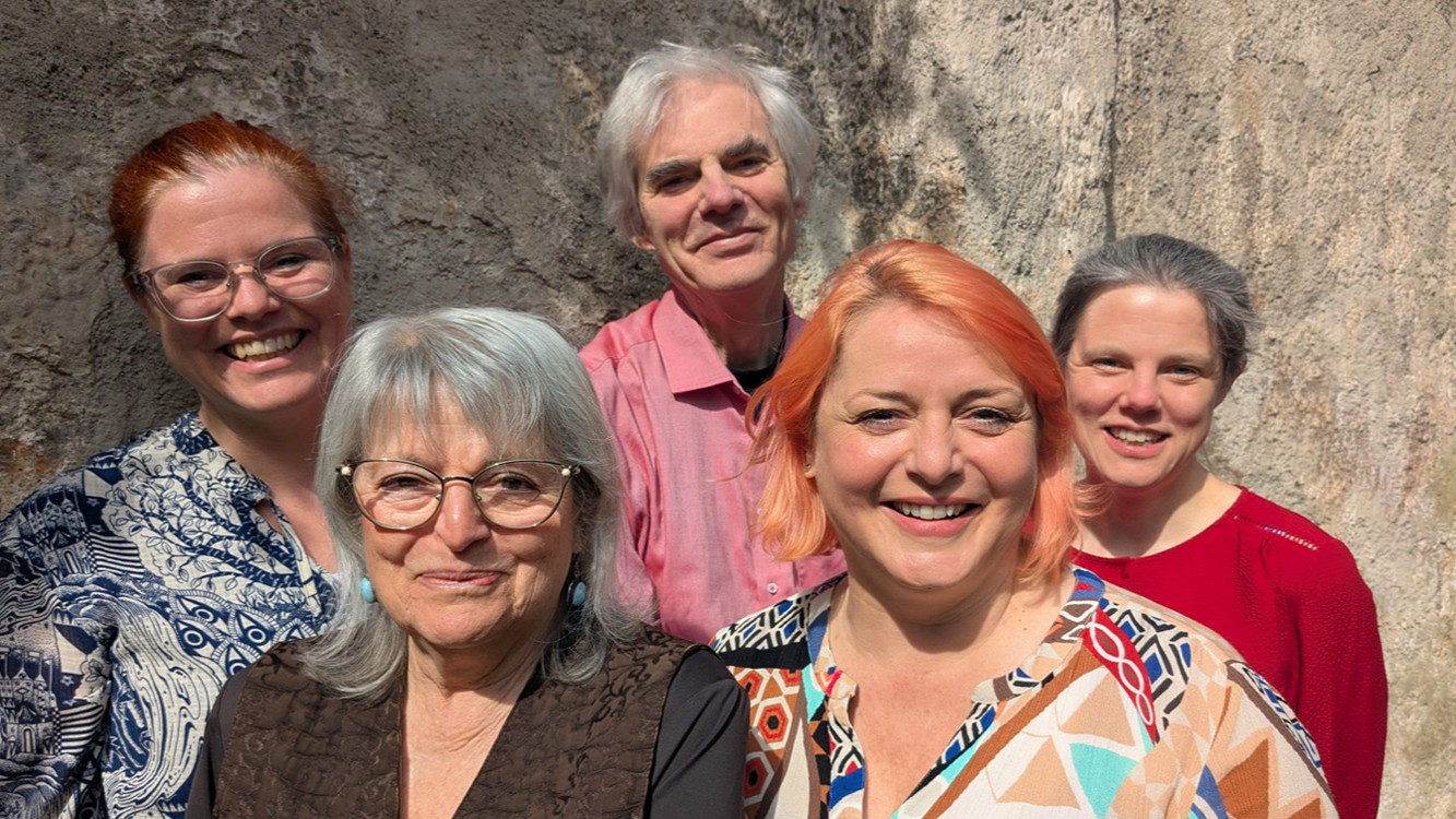 Pressefoto von Weihnachten? Ja bitte! mit fünf Mitwirkenden vor einer Natursteinwand, darunter vier Personen mit Brille und eine Person mit rosa Haaren, in farbenfroher Kleidung und gruppiert für die Kamera.