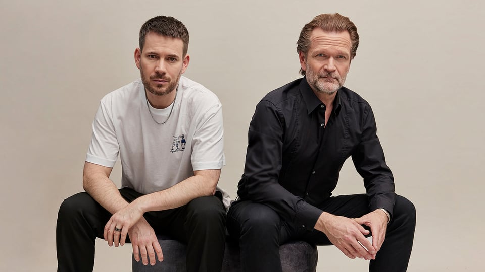 Sebastian Knauer und Johannes Strate sitzen auf einem grauen Hocker vor hellem Hintergrund