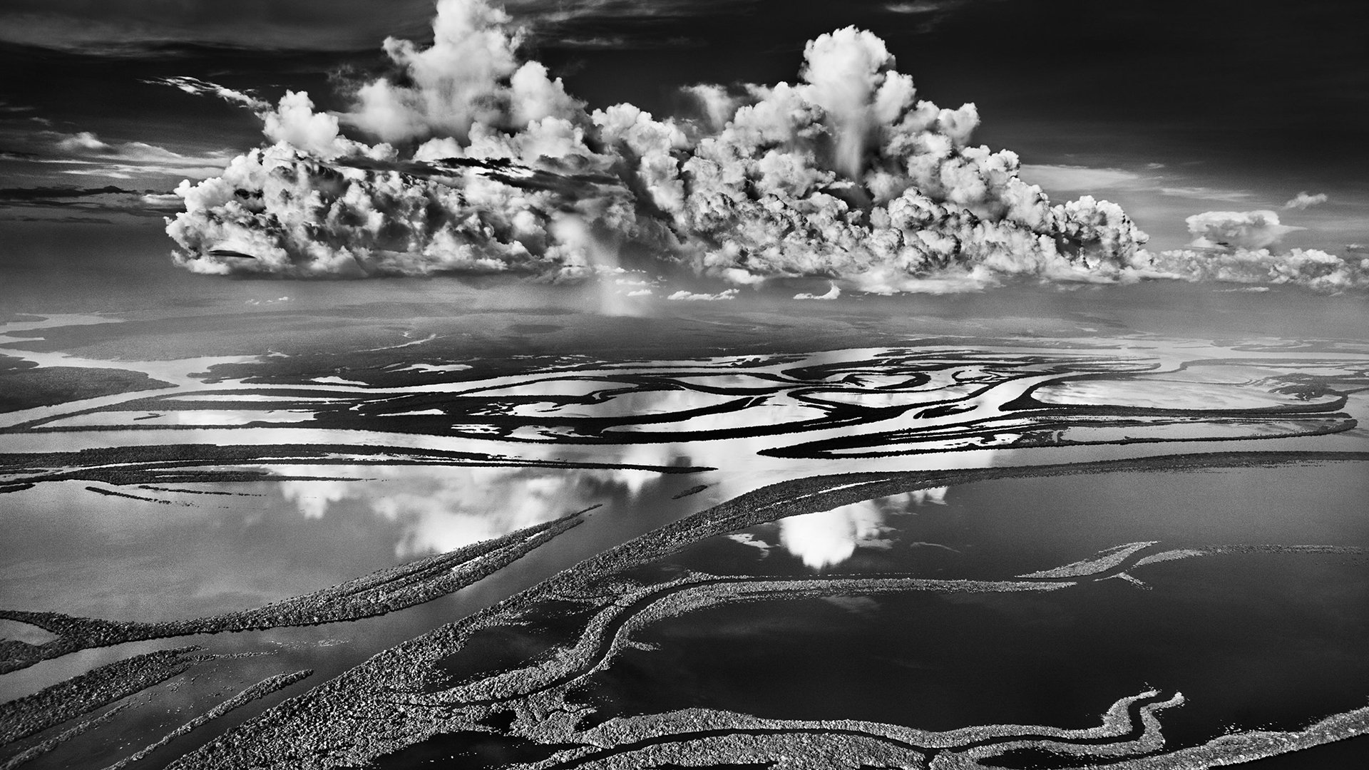 Schwarz-weiß Fotografie einer weitläufigen Flusslandschaft im Amazonas mit verzweigten Wasserläufen, darüber dramatische Wolkenformationen und Spiegelungen im Wasser