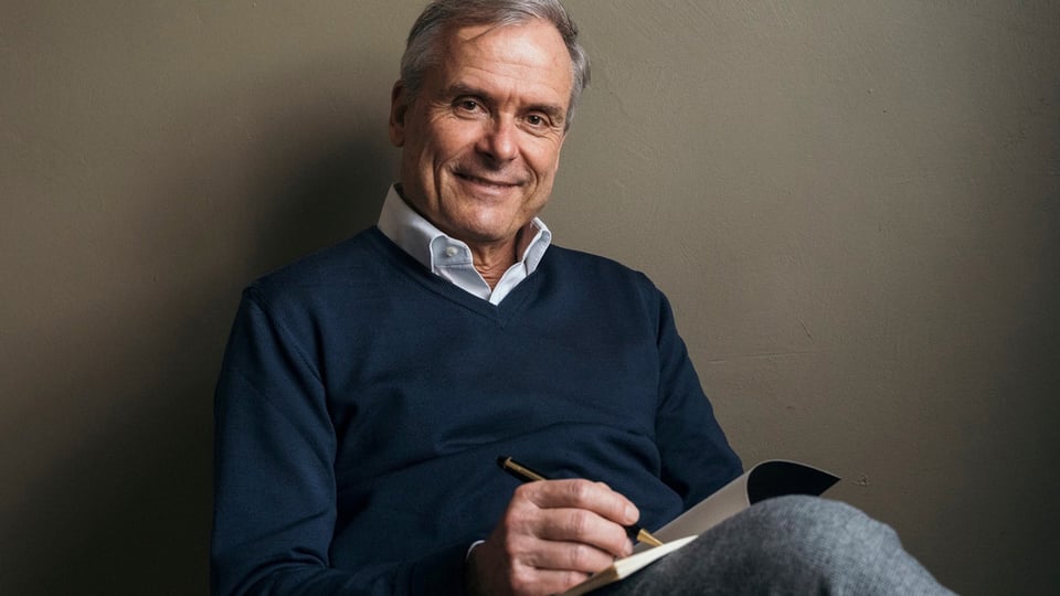 Porträtfoto zu „Axel Hacke – Axel Hacke liest und erzählt“ mit Axel Hacke sitzend, mit Notizbuch und Stift in der Hand, vor neutralem Hintergrund.