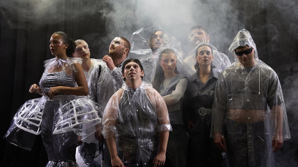 Gruppenaufnahme einer Theaterszene auf einer Bühne. Mehrere Personen stehen dicht beieinander und tragen transparente Regencapes sowie Kostüme in Grau- und Silbertönen. Einzelne Kostüme bestehen aus auffälligen Kunststoff- und Folienelementen. Über der Gruppe liegt dichter Bühnennebel, während dunkle Vorhänge den Hintergrund bilden. Die Inszenierung arbeitet mit Licht, Rauch und ungewöhnlichen Materialien und erzeugt eine atmosphärische, moderne Bühnenästhetik.