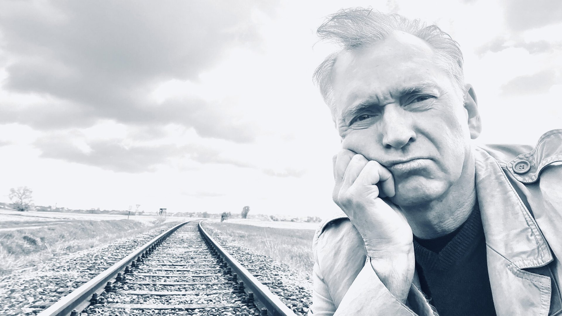 Porträtfoto für eine Veranstaltungsankündigung: Eine Person steht im Vordergrund eines Schwarz‑Weiß‑Motivs und lehnt mit dem Kopf auf der Hand an. Im Hintergrund verlaufen Eisenbahnschienen in die Ferne durch eine offene Landschaft unter bewölktem Himmel. Die Bildkomposition wirkt ruhig und reduziert und eignet sich als Motiv für eine Bühnen‑ oder Kabarettveranstaltung.