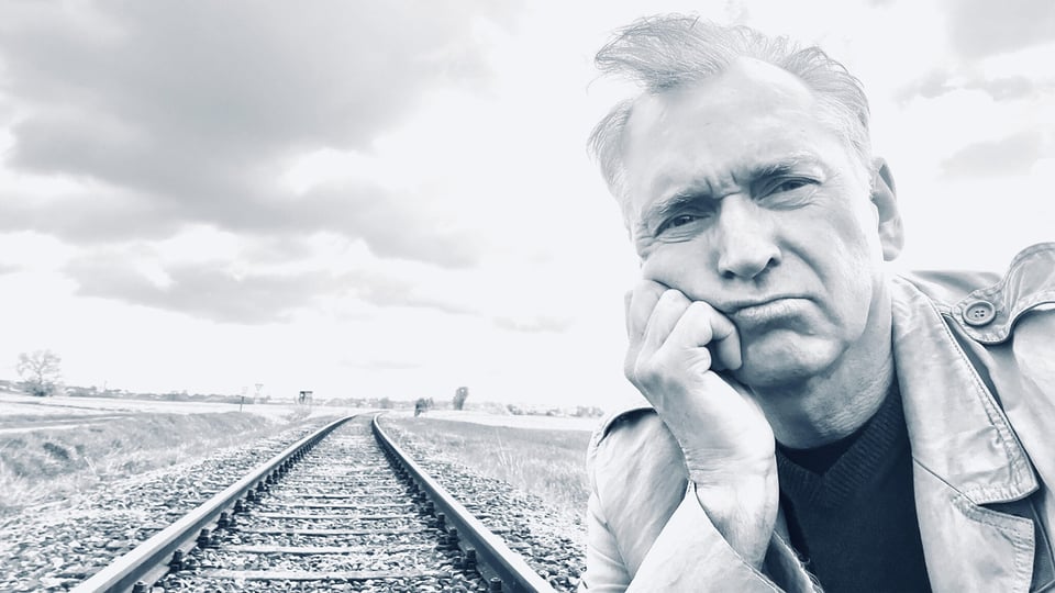 Porträtfoto für eine Veranstaltungsankündigung: Eine Person steht im Vordergrund eines Schwarz‑Weiß‑Motivs und lehnt mit dem Kopf auf der Hand an. Im Hintergrund verlaufen Eisenbahnschienen in die Ferne durch eine offene Landschaft unter bewölktem Himmel. Die Bildkomposition wirkt ruhig und reduziert und eignet sich als Motiv für eine Bühnen‑ oder Kabarettveranstaltung.