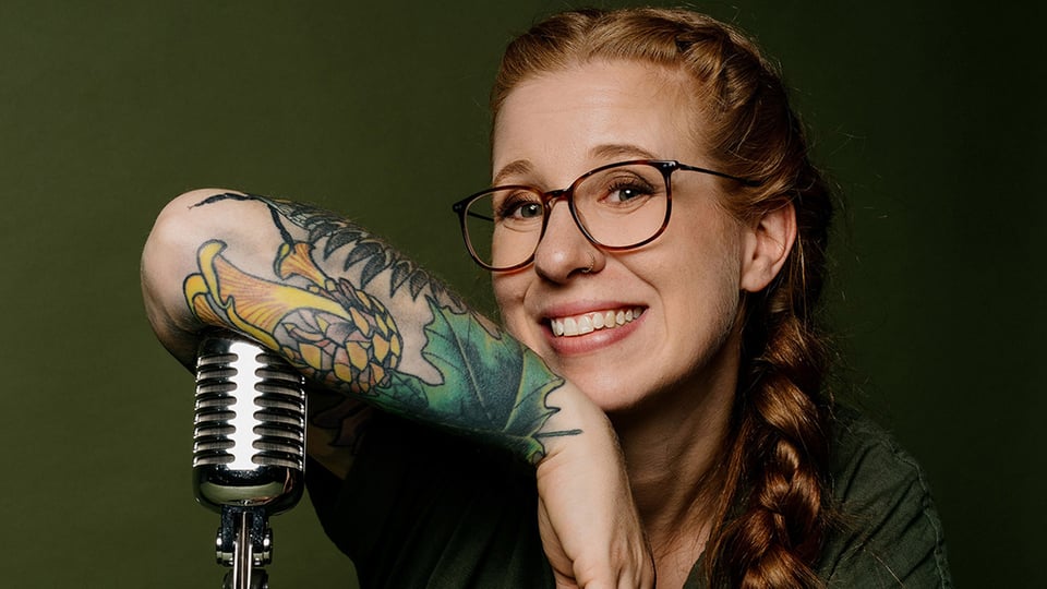Porträtfoto von Sara Brandhuber mit Brille und langem geflochtenem Zopf, die sich auf ein Retro-Mikrofon stützt, vor grünem Hintergrund, mit auffälligem Tattoo am Unterarm.