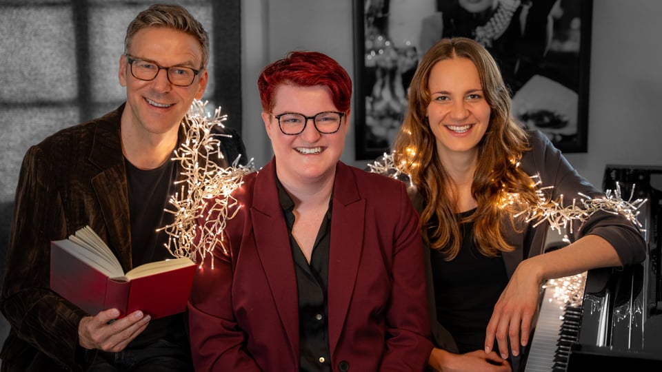 Alexander Netschajew und Duo Key mit drei Personen, Alexander Netschajew hält ein rotes Buch, rechts sitzt eine Pianistin am Klavier. In der Mitte eine Person in dunkelrotem Blazer, eine Lichterkette verläuft durch das Bild, im Hintergrund hängt ein schwarz-weiß Bild
