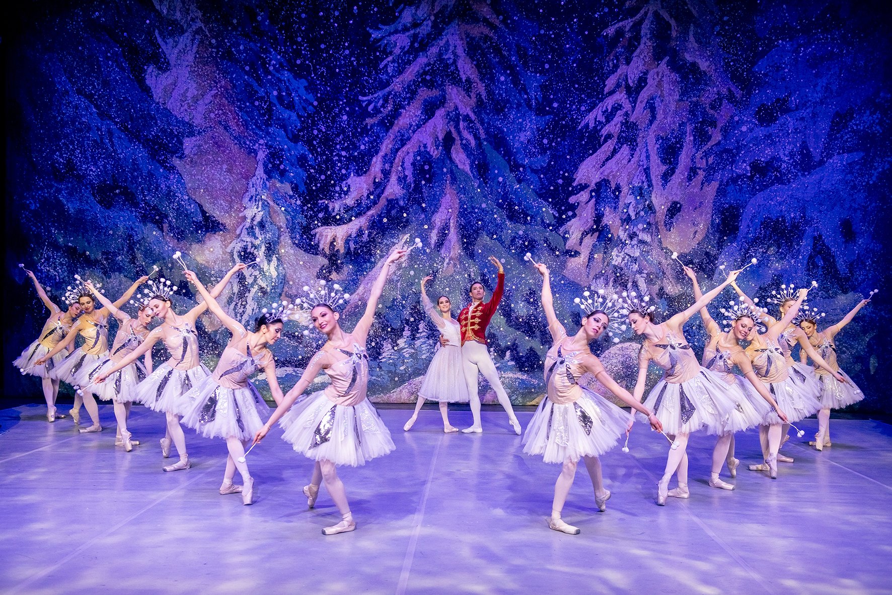 Bühnenfoto aus Der Nussknacker mit einer Solotänzerin und einem Tänzer im roten Kostüm im Zentrum, umgeben von Balletttänzerinnen in weißen Tutu-Kostümen auf einer winterlich gestalteten Bühne vor verschneiter Waldkulisse in Blau- und Violetttönen.