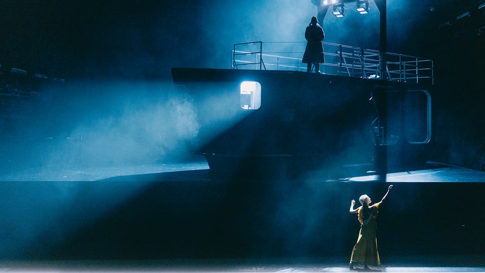 Theaterszene mit schiffsähnlichem Bühnenbild in blauem Licht. Eine Person steht auf dem oberen Deck vor einem Geländer, während eine weitere Person unterhalb auf der Bühne im Lichtkegel in gelben Kleid die Arme erhebt.