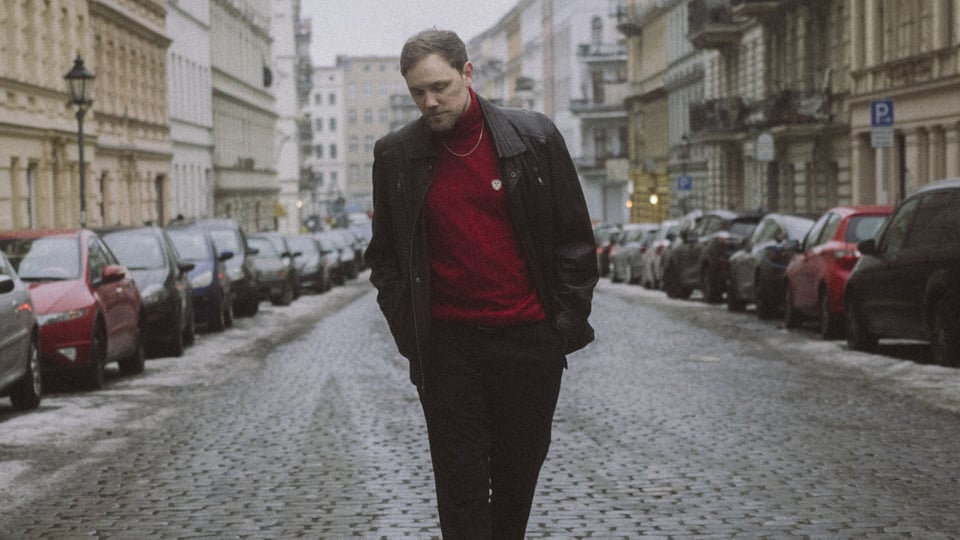 Der Künstler Juse Ju auf einer Straße mit Kopfsteinpflaster, links und rechts ältere Gebäude und parkende Autos