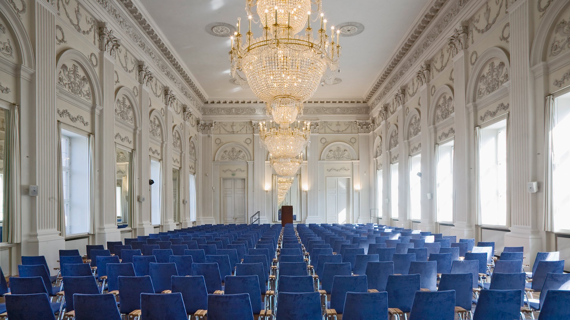 Der Max Joseph-Saal der Residenz mit großem Kronleuchter, Stuckverzierungen an den Wänden, im Vordergrund blaue Besucherstühle