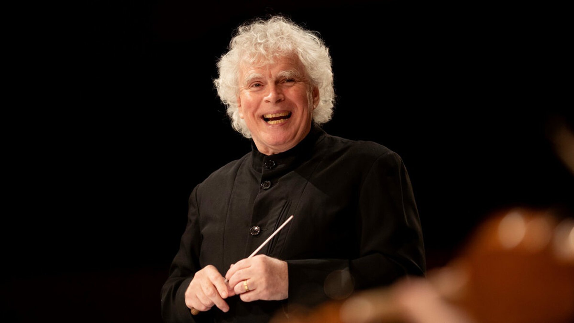 Sir Simon Rattle lachend mit Dirigentenstock vor schwarzem Hintergrund