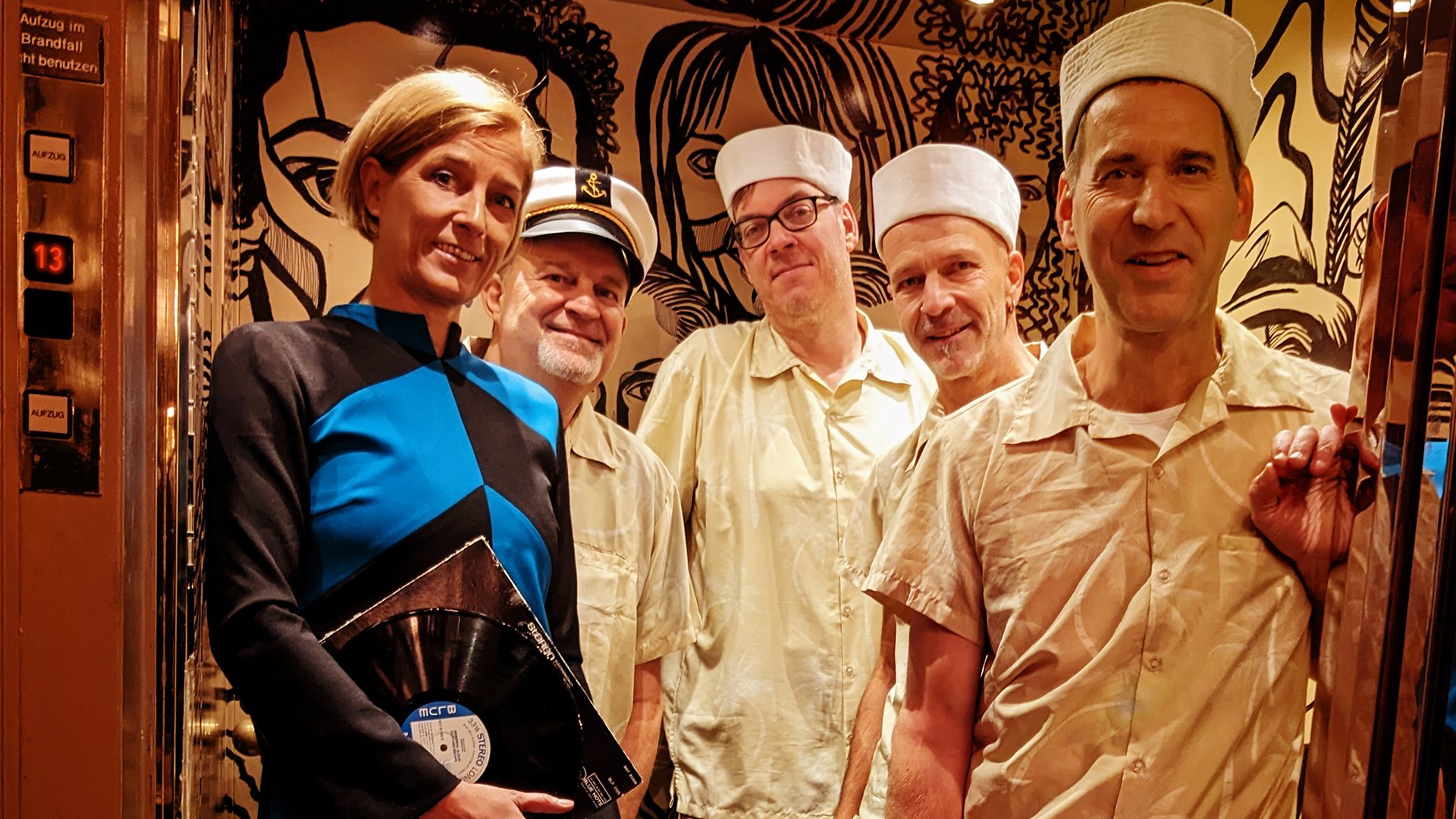 Fünf Mitglieder der Band The Ukelites stehen in einem Aufzug. Eine Person trägt ein blau-schwarzes Outfit und hält eine Schallplatte, vier Personen tragen hellbeige Kleidung mit weißen Mützen. Der Aufzug ist in warmen Braun- und Goldtönen gestaltet und mit großflächigen schwarzen Illustrationen an den Wänden dekoriert.