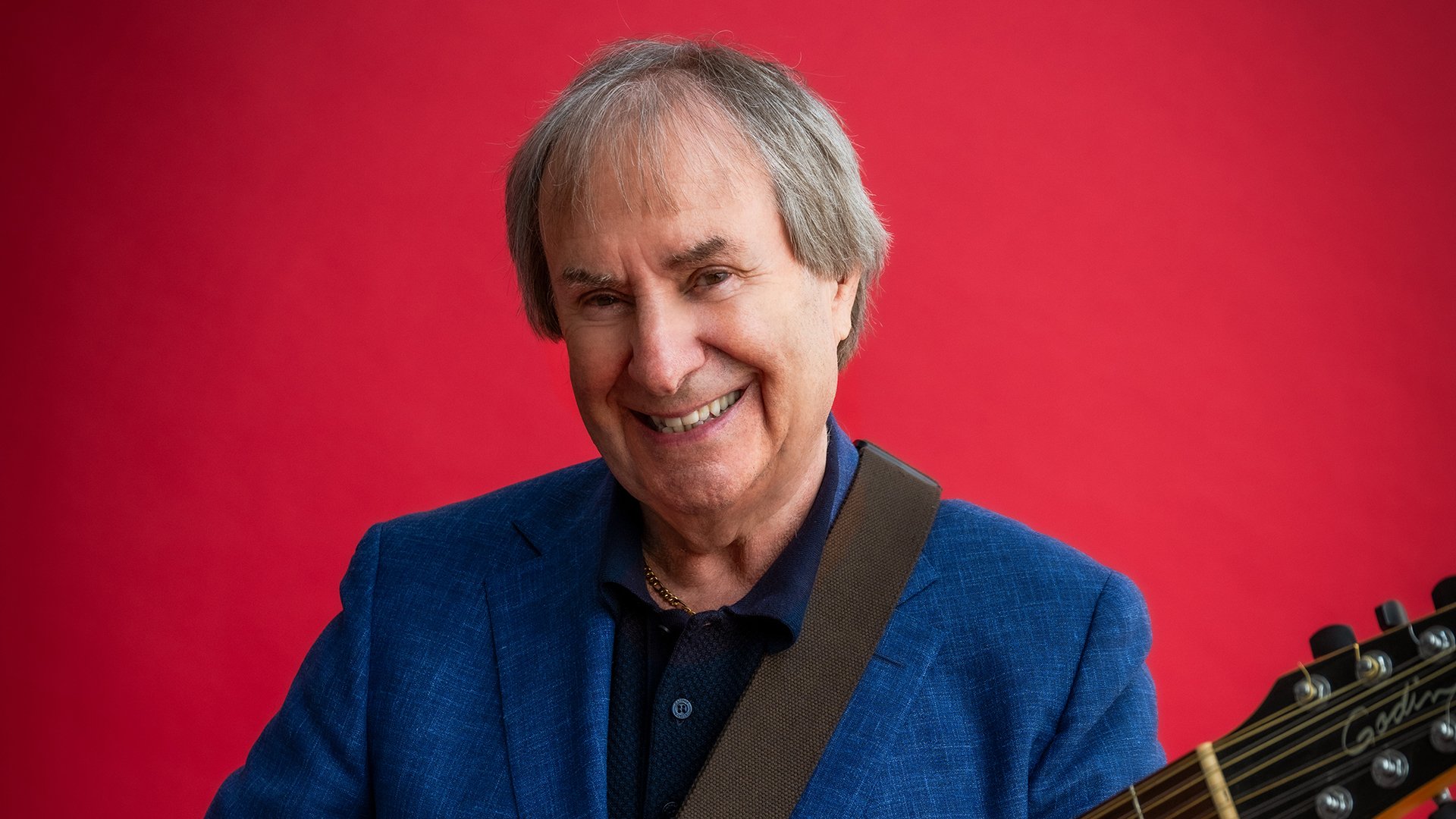 Chris de Burgh lachend mit Gitarre in einem blauen Sakko vor rotem Hintergrund