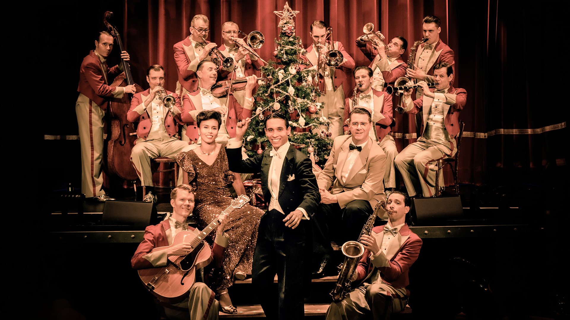 Swing-Ensemble in roten Anzügen spielt vor geschmücktem Weihnachtsbaum auf beleuchteter Bühne
