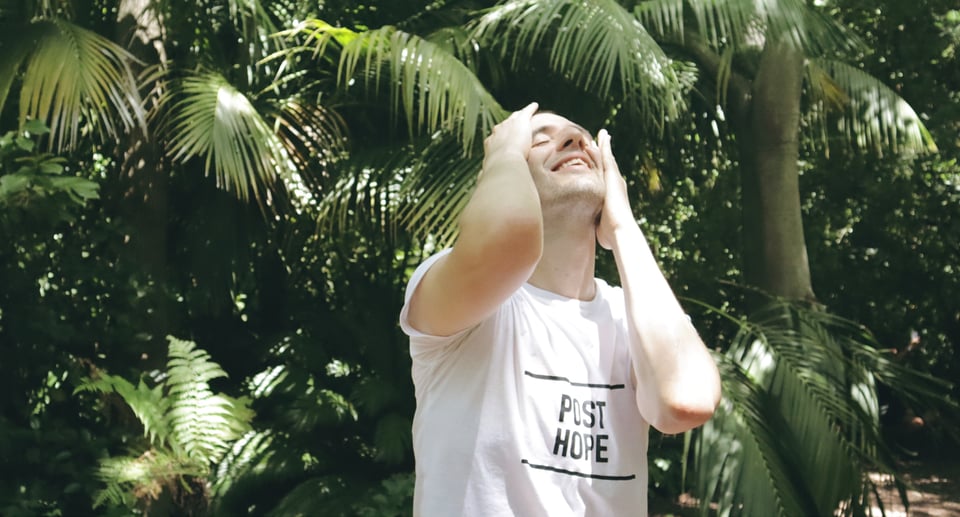Luc Spada steht mit T-shirt vor Palmen in der Sonne und fasst sich durch die Haare und schaut mit geschlossenen Augen in den Himmel nach oben