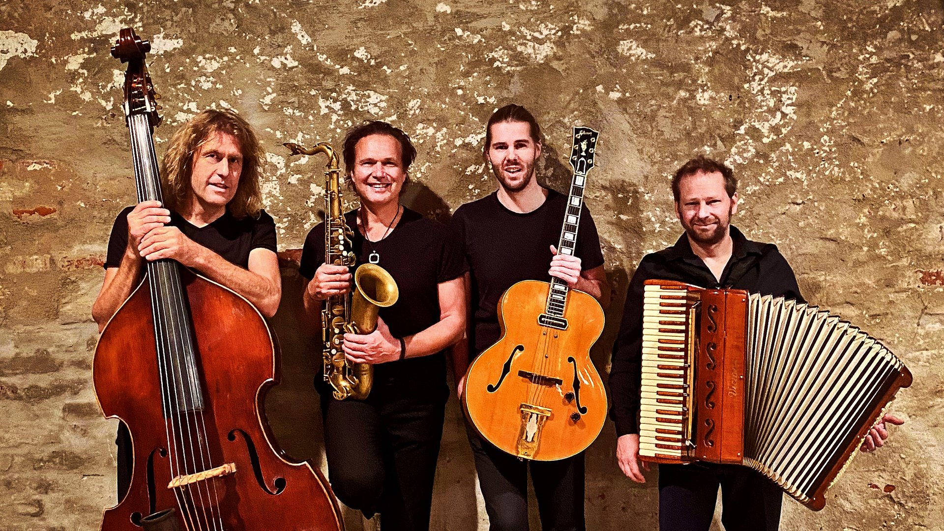 Vier Musiker mit Kontrabass, Saxofon, Gitarre und Akkordeon stehen in schwarzer Kleidung vor einer strukturierten Steinwand und halten ihre Instrumente in die Kamera.