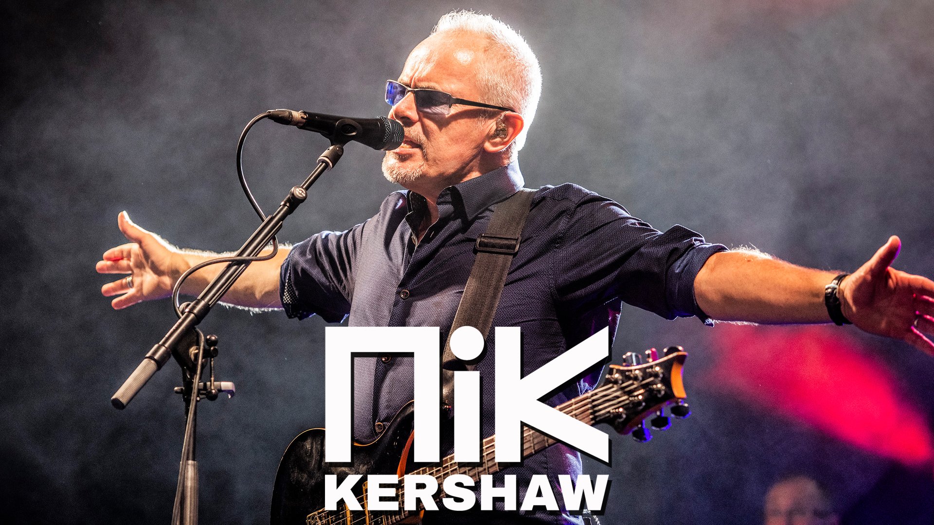 Eventmotiv mit Bühnenaufnahme: Eine Person steht mit ausgebreiteten Armen vor einem Mikrofon und trägt eine E‑Gitarre; Bühnennebel und farbiges Konzertlicht im Hintergrund. Unten im Bild ist der Schriftzug „Nik Kershaw“ zu sehen. Konzert‑Promomotiv für eine Live‑Musikveranstaltung.