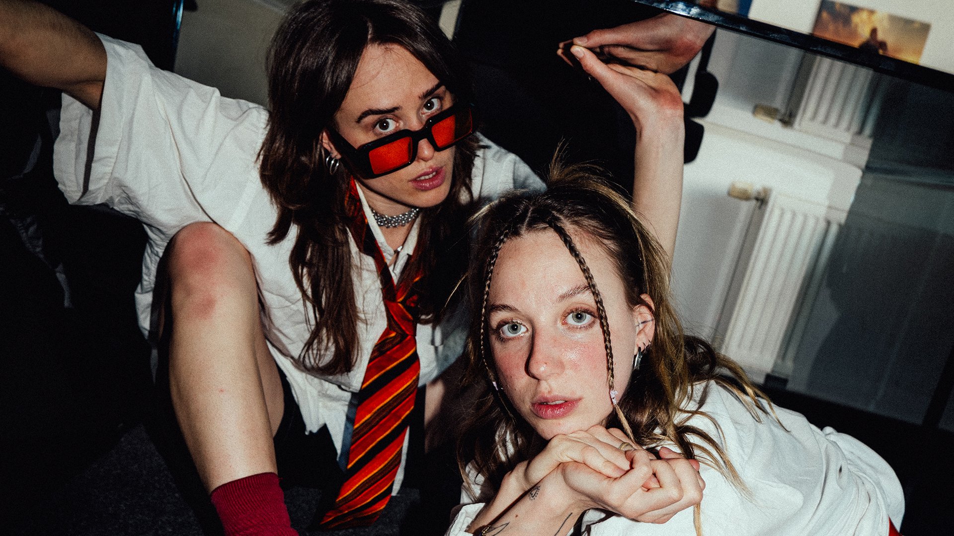 Zwei Personen mit langen Haaren in weißen Hemden, eine mit Krawatte und Sonnebrille