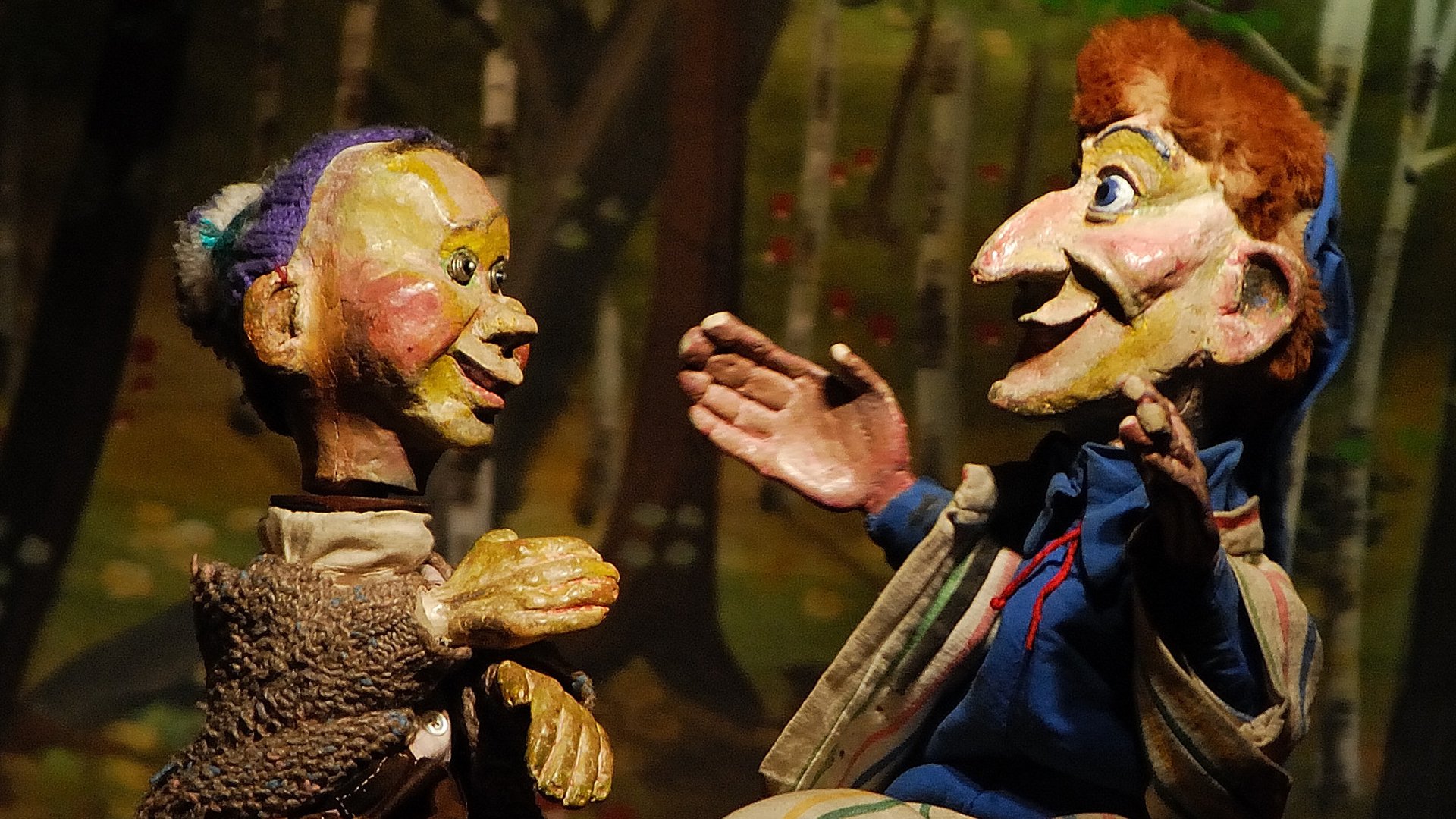 Kasperltheater-Szene mit zwei handgeführten Holzpuppen, die sich gegenüberstehen und miteinander sprechen; detailreich bemalte Figuren mit überzeichneten Gesichtern und Kostümen auf einer Bühnenkulisse im Hintergrund.