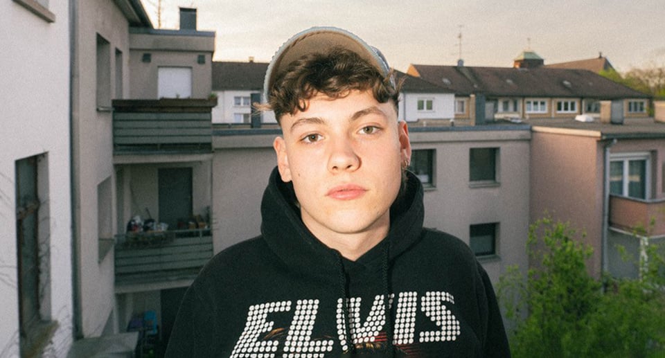 Junger Mann mit Kappe und schwarzem "Elvis"-Sweatshirt vor Hochhauskulisse