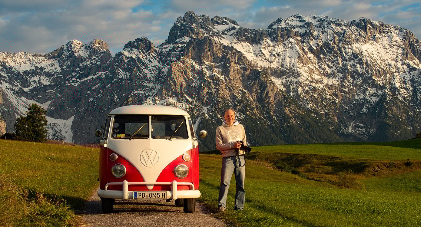 Mann in Jeans auf Wiese stehend eben VW Bully, im Hintergudn Alpen mit Schnee