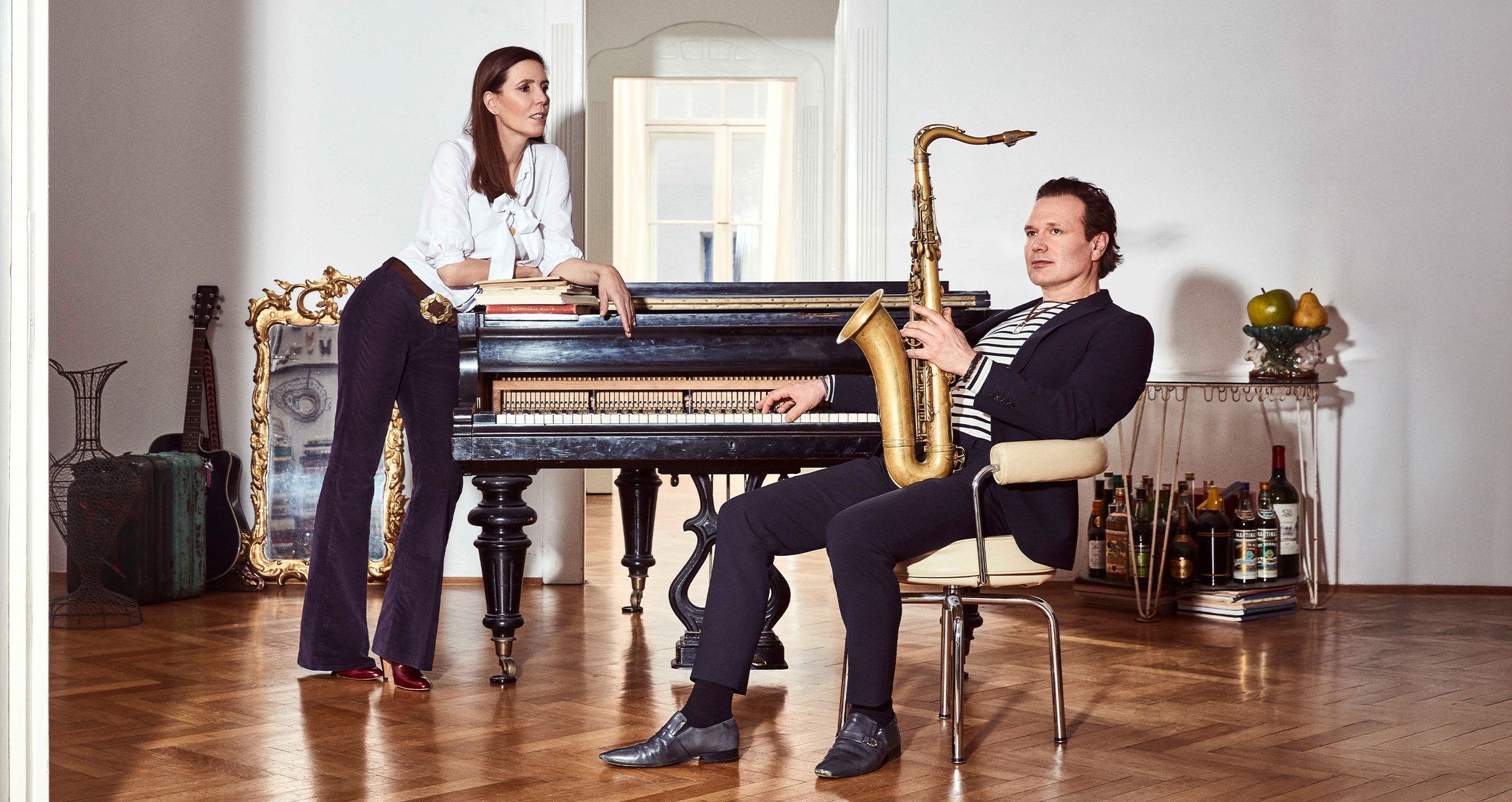 Ein Mann sitzt auf einem Stuhl mit einem Instrument in der Hand und eine Frau lehnt an einem Klavier.