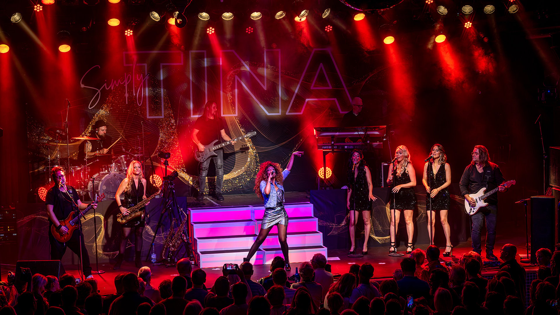 Bühnenaufnahme der Show Simply Tina: Sängerin im Mittelpunkt, Band und Backgroundsängerinnen auf mehreren Ebenen, rotes Bühnenlicht und Publikum im Vordergrund