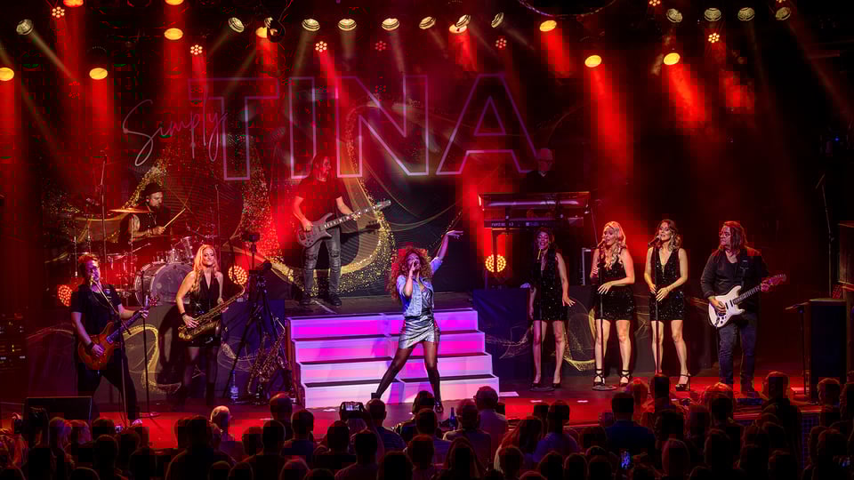 Bühnenaufnahme der Show Simply Tina: Sängerin im Mittelpunkt, Band und Backgroundsängerinnen auf mehreren Ebenen, rotes Bühnenlicht und Publikum im Vordergrund