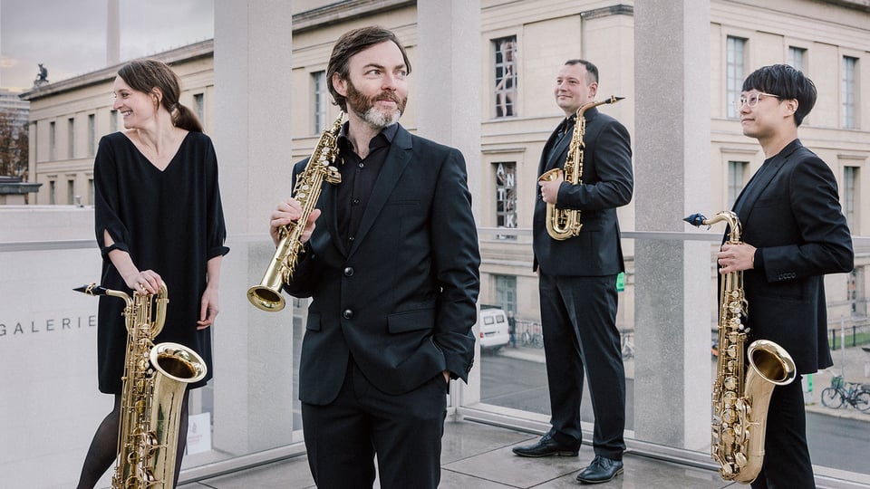 Foto für das Event sonic.art Saxophonquartett, mit der Gruppe ganz in Schwarz gekleidet, mit ihren Blasinstrumenten und einem Gebäude im Hintergrund