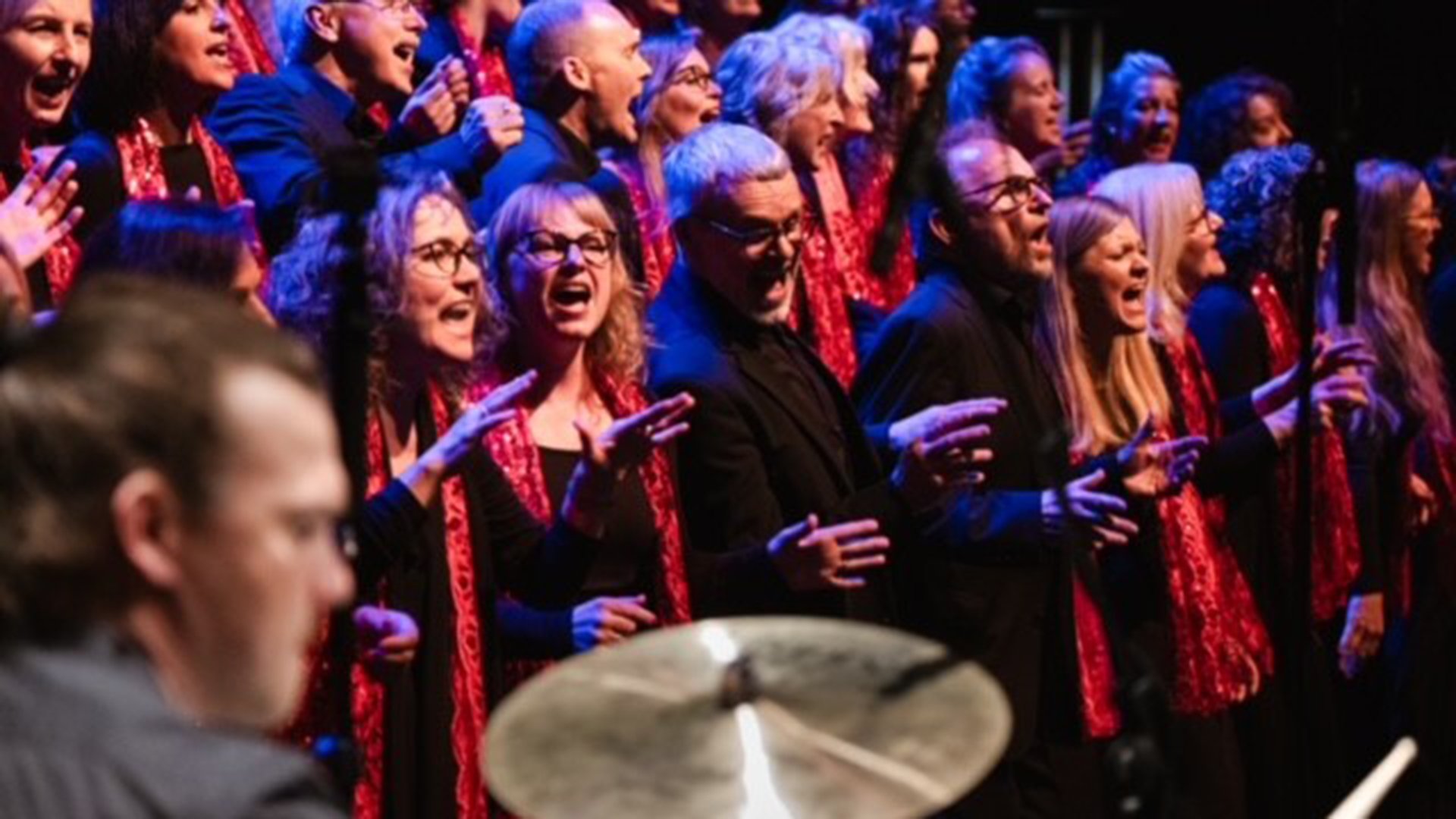 Gospelchor auf einer Bühne während eines Konzerts, im Vordergrund ein Schlagzeug, im Hintergrund singende Chormitglieder mit roten Schals