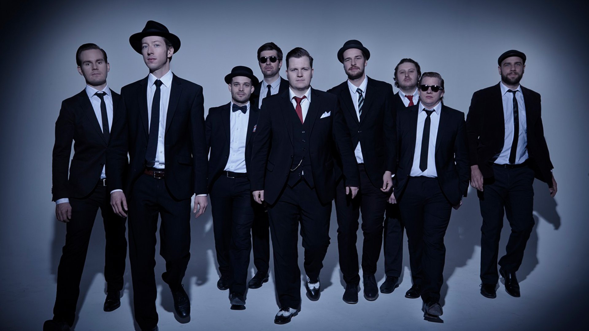 Bandfoto von The Sweet Simones mit neun Personen in dunklen Anzügen und Krawatten, die vor hellem Studiohintergrund auf die Kamera zugehen, einige mit Hut oder Sonnenbrille, eine Person mittig mit roter Krawatte.