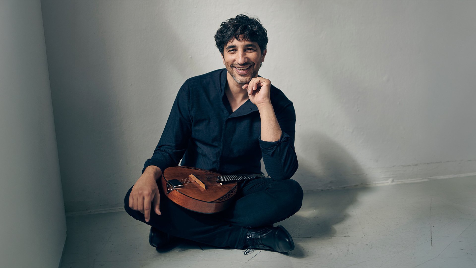 Studioaufnahme einer Person in dunkler Kleidung, sitzend auf dem Boden vor hellem Hintergrund, mit einer Mandoline auf dem Schoß.