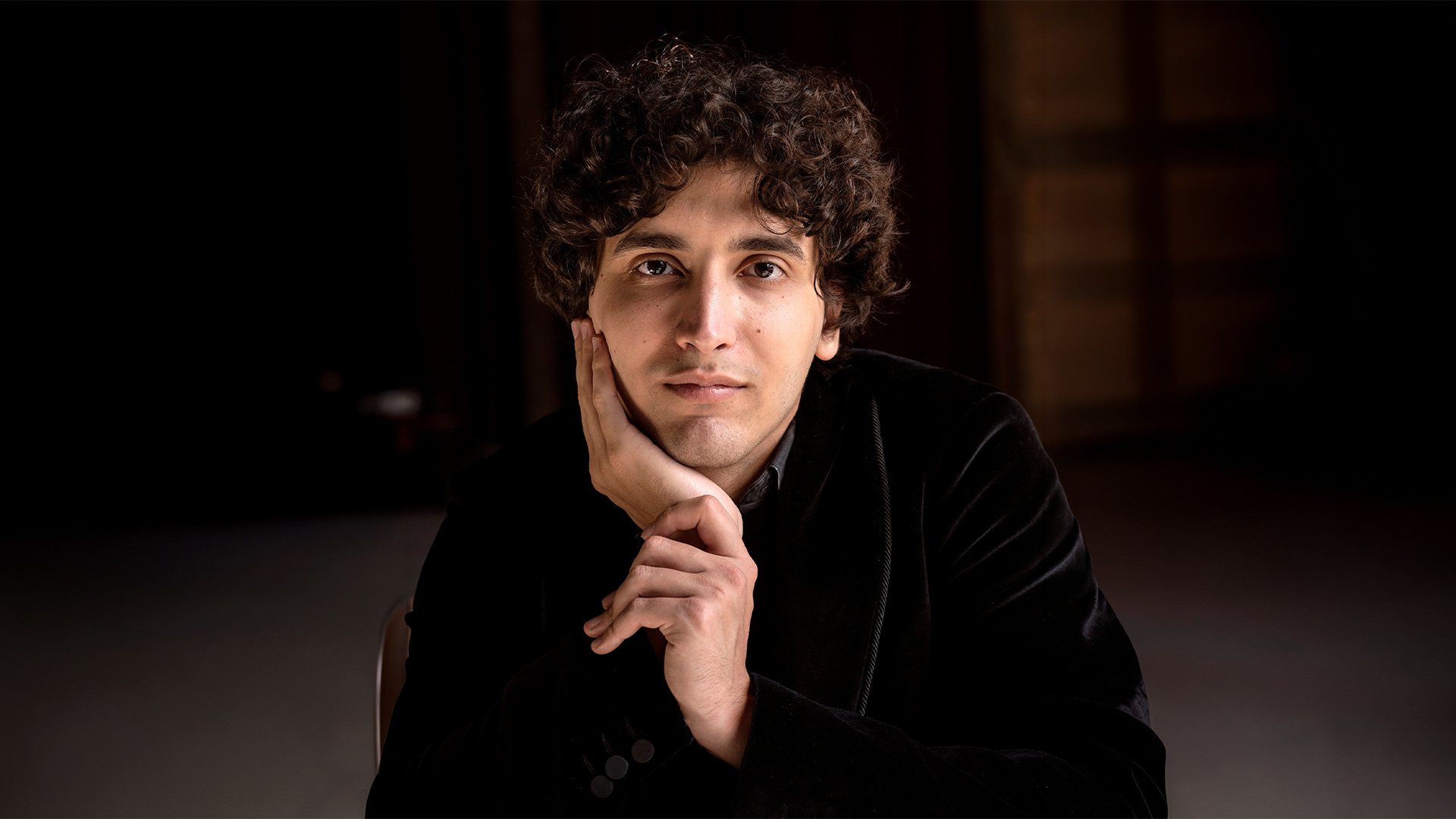 Porträtmotiv für ein klassisches Klavierkonzert: Eine sitzende Person trägt dunkle Kleidung und stützt den Kopf mit einer Hand ab, die andere Hand liegt vor dem Körper. Aufnahme in Innenraum mit geringer Tiefenschärfe; dunkler, unscharfer Hintergrund und weiches, gerichtetes Licht auf Gesicht und Oberkörper. Ruhiges, konzentriertes Promofoto für ein Orchester‑ und Klavierkonzert.
